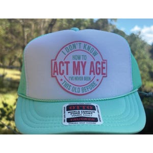 May include: A trucker hat with a white front panel and a mint green brim and mesh back. The front features the text "I DON'T KNOW HOW TO ACT MY AGE I'VE NEVER BEEN THIS OLD BEFORE" in a pink and black design.