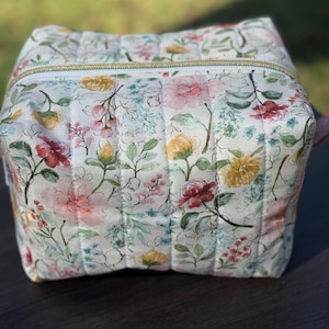 May include: A quilted, floral-patterned cosmetic bag with a gold zipper. The bag features a white background with a repeating pattern of pink, yellow, and red flowers with green leaves. The bag is boxy and sits on a dark surface.