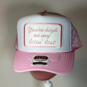 May include: A pink and white trucker hat with the text "You're high on my bitin' list" embroidered on a rectangular patch. The hat features a pink brim and mesh backing, with a "OTTO" label.