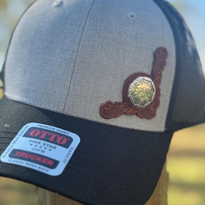 May include: A gray and black trucker hat with a decorative leather patch. The patch features a silver emblem and brown leather accents. The hat has a mesh back and a curved brim. The hat has the text "OTTO" and "TRUCKER" on a sticker.