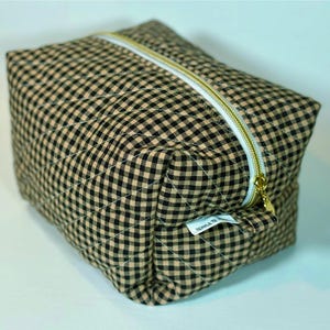 May include: A quilted, rectangular cosmetic bag with a black and tan gingham pattern. The bag features a gold-colored zipper and a small white tag. The stitching creates a diamond pattern across the fabric.