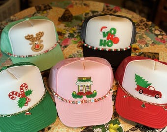 May include: Five Christmas-themed trucker hats with white fronts and mesh backs. Designs include a reindeer, candy canes, elf legs, "HO HO" text, and a truck carrying a Christmas tree. Each hat has a decorative band around the brim.
