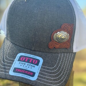 May include: A gray trucker hat with a white mesh back. The hat features a brown leather patch with a silver-colored metal embellishment. The hat has the text "OTTO FIVE STAR TRUCKER" on a blue sticker.