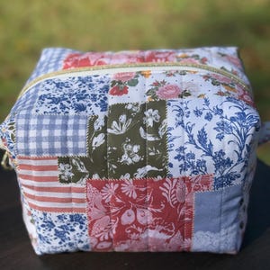 May include: A patchwork fabric box with a gold zipper. The box features a variety of floral and patterned fabrics in shades of red, blue, green, and white. The box is square-shaped and appears to be quilted.