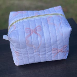 May include: A white quilted cosmetic bag with a repeating pattern of pink bows. The bag has a gold zipper closure and a small fabric loop on the side. The bag is sitting on a dark surface.