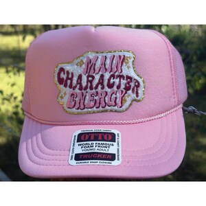 May include: A pink trucker hat with a mesh back and a curved brim. The front of the hat features the text "MAIN CHARACTER ENERGY" in a stylized design. The hat has a snap closure and a soft sweatband.
