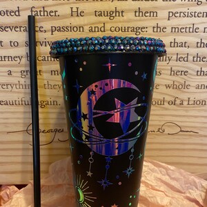 May include: A black reusable cup with a glittery iridescent design of a crescent moon, a star, and other celestial elements. The cup has a black lid with rhinestones around the edge. A black straw is next to the cup.