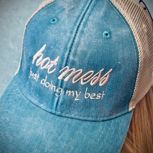 Puede incluir: Una gorra de camionero azul turquesa y beige con un aspecto desgastado. La gorra tiene una parte trasera de malla y una visera curvada. La parte delantera de la gorra tiene el texto "hot mess just doing my best" bordado en un color marrón claro.