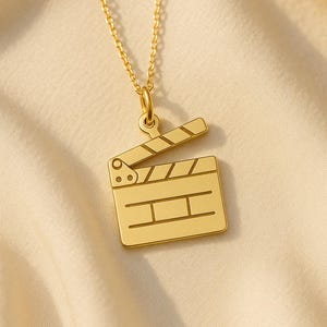 May include: A gold-coloured necklace with a film clapperboard pendant. The pendant has a rectangular base with horizontal lines and a hinged top. The chain is a delicate gold colour, set against a soft, neutral fabric background.
