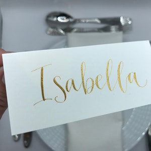 May include: White place card with the name "Isabella" written in gold calligraphy.