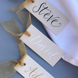May include: Three white place cards with gold ribbon tied around them. The names "Steve", "David", and "Adam" are written in calligraphy on the cards.