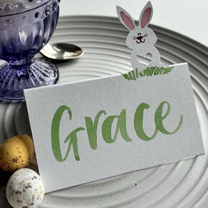 May include: A white card with the name "Grace" in green script sits on a grey plate, next to a paper bunny. A purple glass egg cup holds a white egg. Two speckled eggs are also on the plate, suggesting an Easter theme.