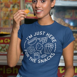 May include: A woman wearing a blue t-shirt with the text "I'm just here for the snacks" and a variety of snack food icons. She is holding a small snack in her right hand and a small bowl in her left hand.