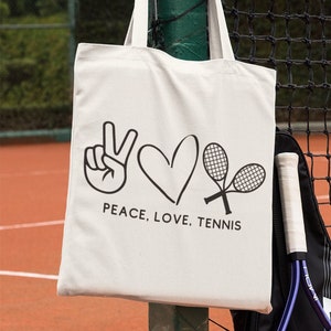 May include: White canvas tote bag with black text that reads "Peace, Love, Tennis" and a peace sign, heart, and tennis racket graphic. The bag is hanging on a tennis net.