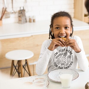 May include: A young person wearing a white long-sleeved shirt with the text "I'm just here for the snacks" and a graphic of various snacks. They are eating a cookie and smiling at the camera.
