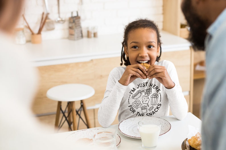 May include: A young person wearing a white long-sleeved shirt with the text "I'm just here for the snacks" and a graphic of various snacks. They are eating a cookie and smiling at the camera.