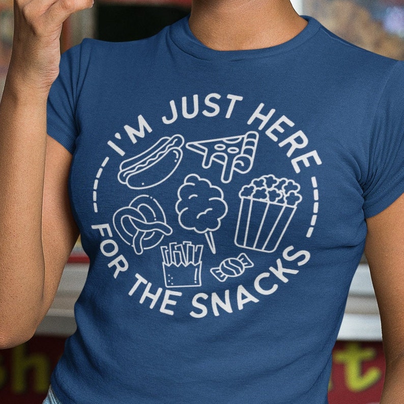 May include: A blue t-shirt with a white graphic design that says "I'm just here for the snacks" and features various snack foods like popcorn, pretzels, hot dogs, pizza, candy, and french fries.