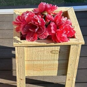 May include: A wooden planter box with a square shape and four legs. The planter is made of natural wood and has a light brown color. The planter is filled with a bouquet of artificial pink peonies.