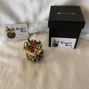 May include: A decorative, jewel-toned Santa's Bag figurine, filled with miniature gifts and a stocking. The bag is navy blue with gold accents and a gold chain. The figurine is accompanied by a black box and a tag that reads "Santa's Bag".