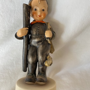 May include: A ceramic figurine of a chimney sweep boy wearing a black and brown outfit, holding a long black brush and a bag over his shoulder. He is standing on a cream-coloured base.