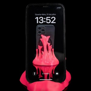 May include: A pink 3D printed phone stand with a textured surface. The stand is holding a black smartphone with a bright pink screen displaying the time 13:52 and the date "Quarta-feira, 31 de julho".