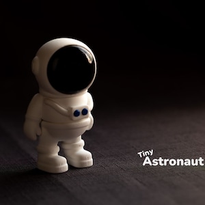 Puede incluir: Una figurita de astronauta de plástico blanco con una visera negra y detalles azules. La figurita está de pie sobre una superficie marrón oscura. El texto "Tiny Astronaut" está escrito en blanco debajo de la figurita.