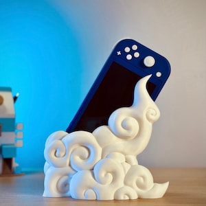 Puede incluir: Soporte en forma de nube de color blanco impreso en 3D con una consola Nintendo Switch azul apoyada en él.