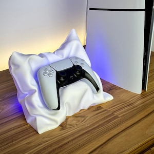 May include: A white PlayStation 5 console with a white PlayStation 5 DualSense controller resting on a white pillow-shaped stand. The stand has a blue light emanating from the bottom.