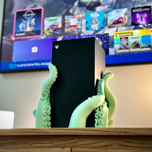 Könnte beinhalten: Eine schwarze Spielekonsole mit einem grünen, krakenförmigen Ständer. Die Konsole steht auf einer Holzoberfläche. Die Konsole hat ein Logo auf der Vorderseite.