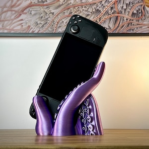 Pode incluir: Um suporte de polvo impresso em 3D roxo segurando um dispositivo de jogo portátil preto. O suporte tem uma superfície texturizada e o dispositivo tem um botão circular na parte superior.