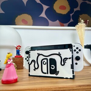 May include: White Nintendo Switch console with a black and white doodle design on the stand. The console is on a wooden surface with a small pink princess figurine and a red and blue Mario figurine.