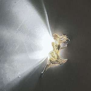 May include: A golden sculpture of a woman with flowing hair, illuminated by a bright light source, creating a dramatic effect against a white textured background.