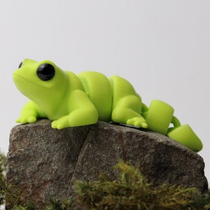Könnte beinhalten: Eine kleine, leuchtend grüne, handgefertigte Eidechsenfigur mit schwarzen Augen, die auf einem mit Moos bewachsenen Stein sitzt. Die Eidechse ist in einer Pose mit nach oben geneigtem Kopf und um den Stein gewickeltem Schwanz dargestellt. Der Text "Baby Lizard" ist auf dem Bild zu sehen.