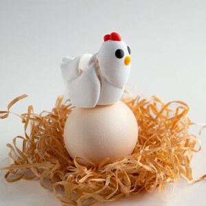 Può includere: Una statuetta di pollo in ceramica bianca posata su un uovo bianco, annidato in un letto di paglia marrone. Il testo "Tiny Chicken" è scritto sotto l'uovo.