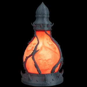 May include: A gray, gothic-style lamp with a skull design. The lamp has a pointed top and is encircled by thorny vines. The interior glows with an orange light, and the base has a series of spikes.
