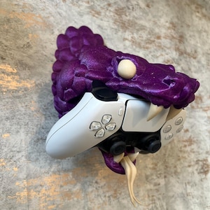 Peut inclure: Un support mural en forme de tête de dragon imprimé en 3D de couleur violette avec une manette de jeu vidéo blanche et noire. Le dragon a une surface texturée et un œil blanc.
