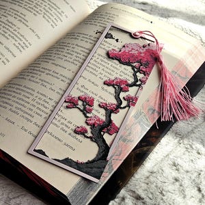 K&ouml;nnte beinhalten: Dekorativer Lesezeichen mit einem Baumdesign, das rosa Bl&uuml;ten und einen schwarzen Stamm aufweist. Das Lesezeichen hat eine rosa Quaste und befindet sich in einem offenen Buch. Das Lesezeichen ist aus Papier.