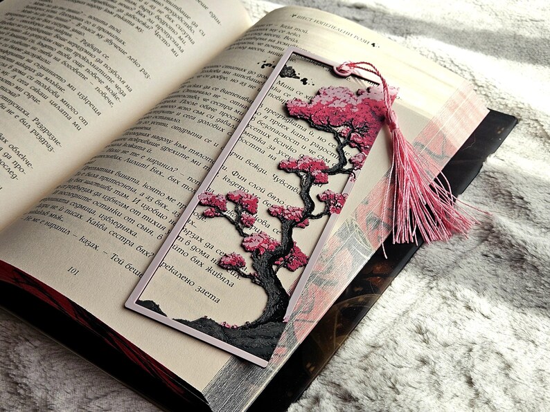 K&ouml;nnte beinhalten: Dekorativer Lesezeichen mit einem Baumdesign, das rosa Bl&uuml;ten und einen schwarzen Stamm aufweist. Das Lesezeichen hat eine rosa Quaste und befindet sich in einem offenen Buch. Das Lesezeichen ist aus Papier.
