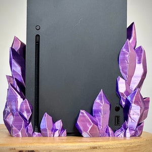 May include: A black gaming console with a purple crystal-shaped stand. The stand is made of 3D printed plastic and has a glossy finish.