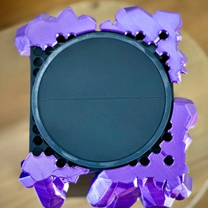 May include: A purple 3D printed crystal-shaped stand with a black circular base. The stand has a textured surface and is designed to hold a small object.