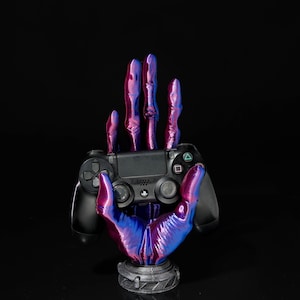 May include: A black PlayStation controller held in a 3D printed hand with a purple and blue iridescent finish. The hand is on a black base.