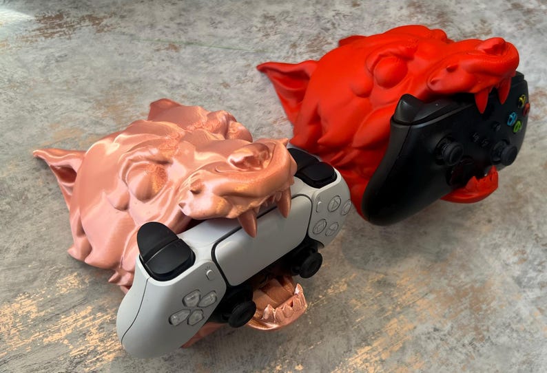 May include: Two 3D printed wolf head game controller holders. One is a red wolf head holding a black Xbox controller. The other is a copper wolf head holding a white Playstation controller.