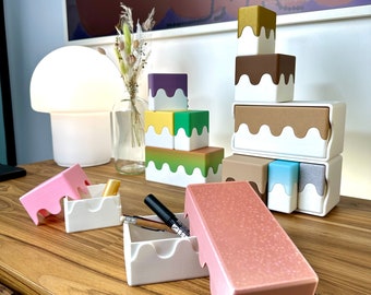 May include: A set of colorful, stackable storage boxes with a unique drip design. The boxes are made of white plastic with a variety of colors on the top. The boxes are arranged in a stack on a wooden table.
