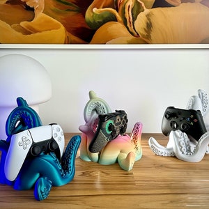 Puede incluir: Tres soportes para mandos de videojuegos con forma de pulpo en color turquesa, arcoíris y blanco. Cada soporte sujeta un mando de juego diferente, incluyendo un mando blanco de PlayStation 5 y un mando negro de Xbox. Los soportes están sobre una superficie de madera.