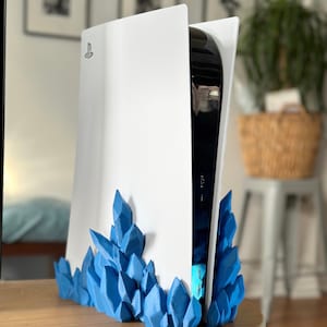 Op de afbeelding: Een witte PlayStation 5 console met een blauwe kristalvormige standaard. De console staat rechtop en de standaard bevindt zich aan de onderkant van de console.