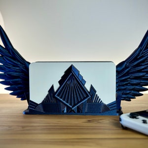 Può includere: Un supporto per schede di gioco Nintendo Switch stampato in 3D in bianco e nero con ali. Il supporto ha un design geometrico e si trova su una superficie in legno.