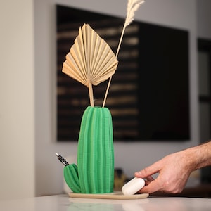 May include: A green cactus-shaped vase with a pen in a side pocket and a beige tray. The vase holds a dried fan-shaped leaf and a tall, thin, feathery plant stem. A hand reaches for a white, rectangular object on the tray.