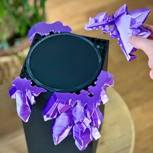 May include: A black speaker with a purple crystal-shaped design around the top. The crystal design is removable and is being held in a hand.