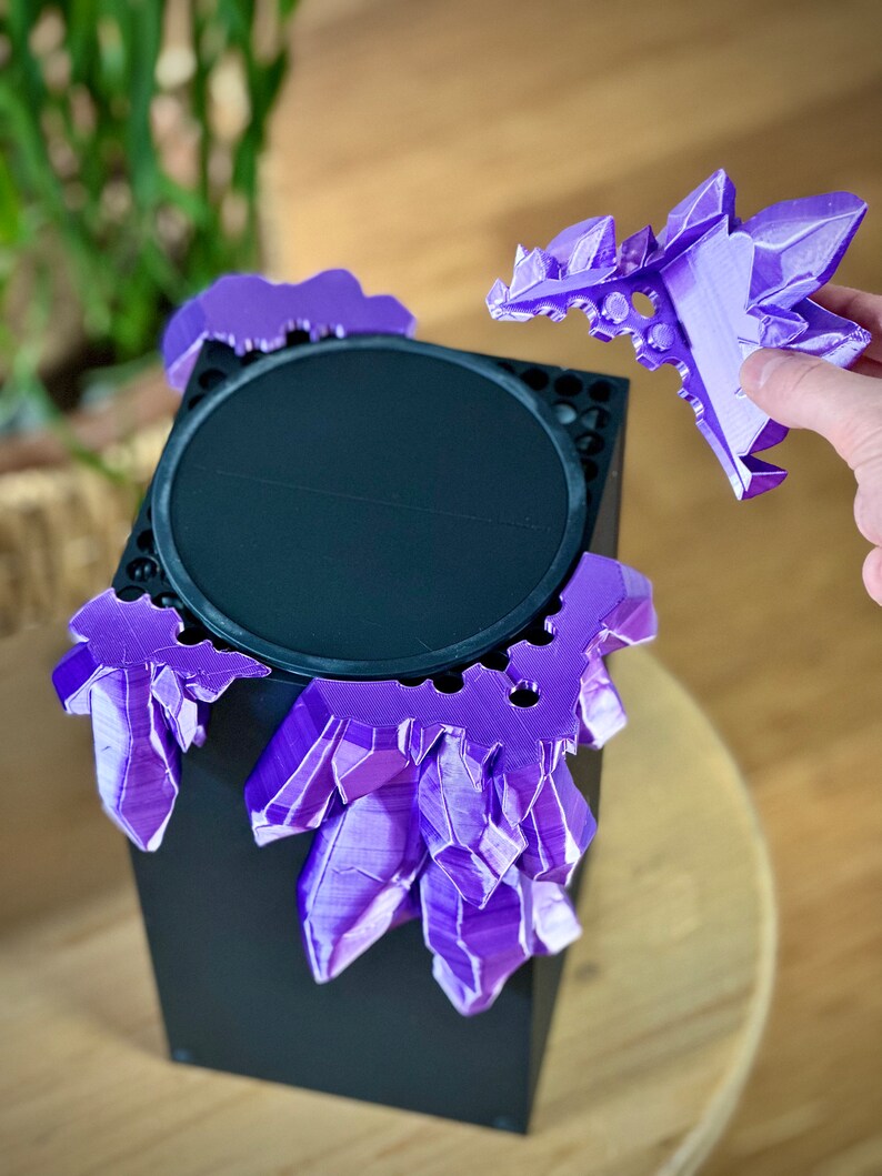 May include: A black speaker with a purple crystal-shaped design around the top. The crystal design is removable and is being held in a hand.
