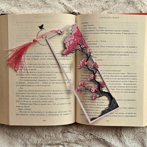 K&ouml;nnte beinhalten: Transparenter Acryl-Lesezeichen mit einem Kirschbl&uuml;ten-Design mit rosa Bl&uuml;ten und einem schwarzen Baumstamm. Das Lesezeichen hat eine rosa Quaste und liegt in einem offenen Buch.
